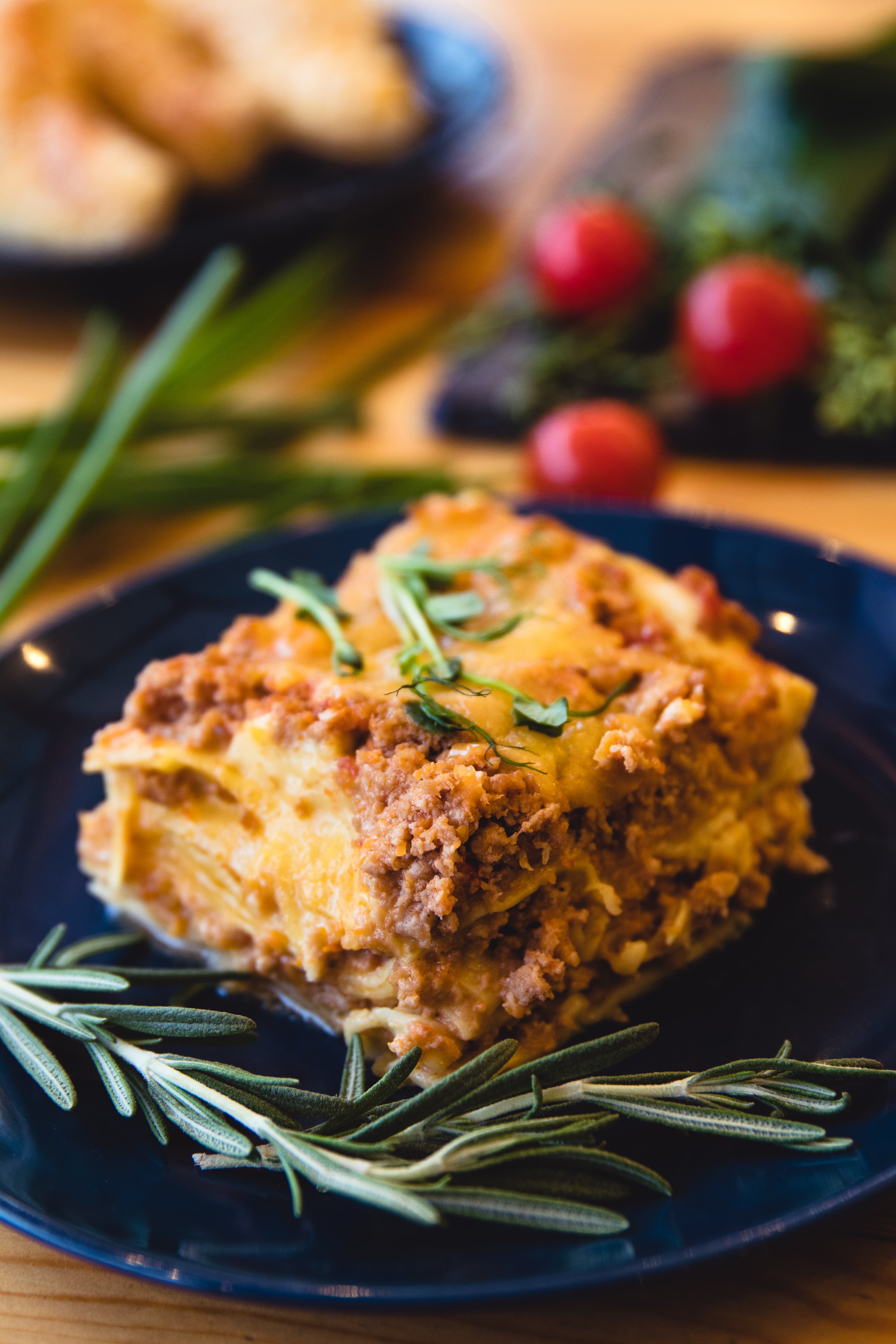 A slice of layered lasagna on a dark blue plate, garnished with fresh rosemary, with cherry tomatoes and herbs behind it
